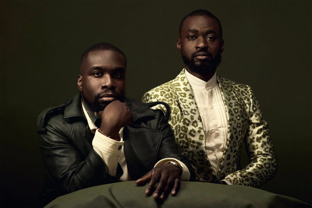 Two men pose against a dark background; one wears a black leather jacket and rests his chin on his hand, while the other stands beside him in a white shirt and a green leopard-print blazer.