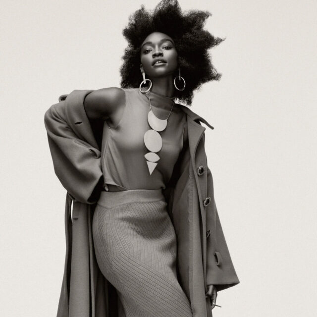 A stylish woman with natural hair poses in a long coat over a sleeveless top and ribbed skirt. She wears bold geometric jewelry, including large earrings and a statement necklace, against a simple background.