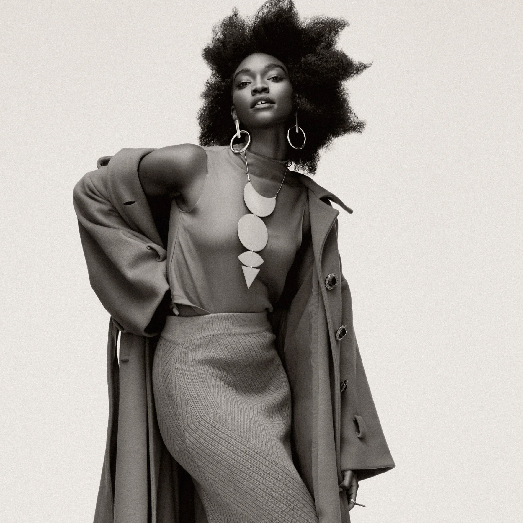 A stylish woman with natural hair poses in a long coat over a sleeveless top and ribbed skirt. She wears bold geometric jewelry, including large earrings and a statement necklace, against a simple background.