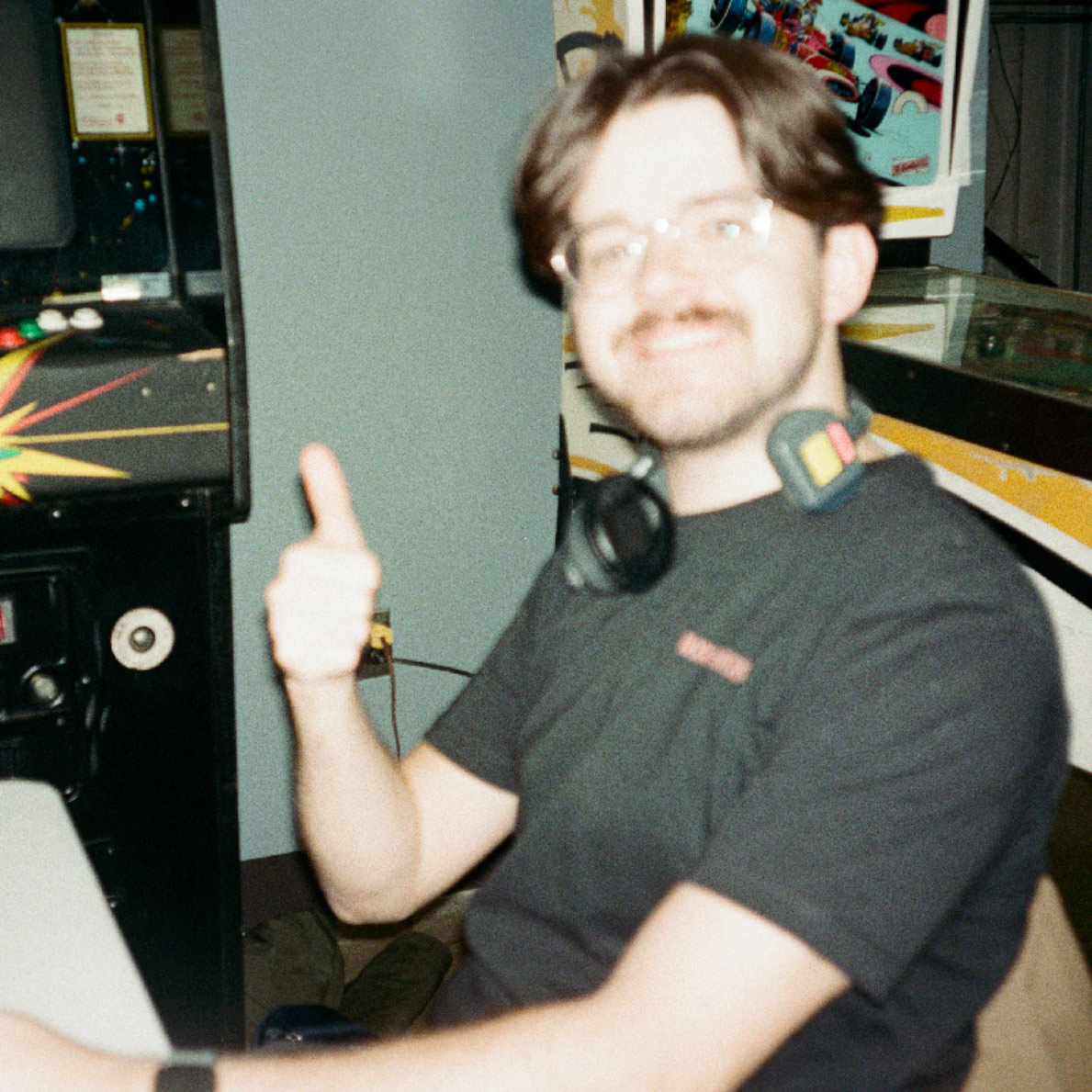 Director, Nathan Colby, smiles and gives a thumbs up while sitting at an arcade machine. They are wearing headphones around their neck and a black shirt. The image is slightly blurry.
