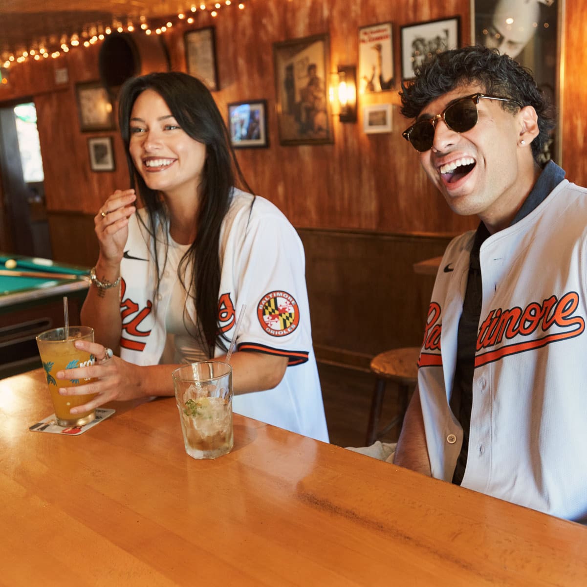 Two people wearing Baltimore Orioles jerseys sit at a bar, smiling and enjoying drinks. The bar has wooden walls, framed photos, string lights, and a pool table in the background.
