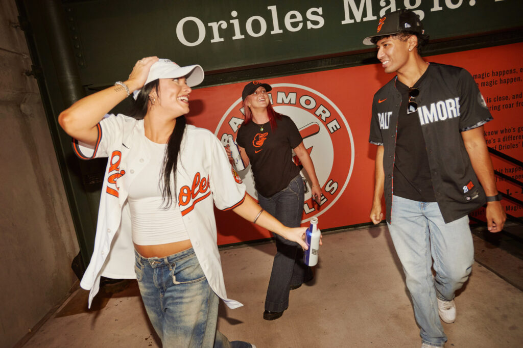 Three young adults wearing Baltimore Orioles baseball jerseys and caps smile and walk together under a sign reading “Orioles Magic” at a stadium, with one person holding a drink.