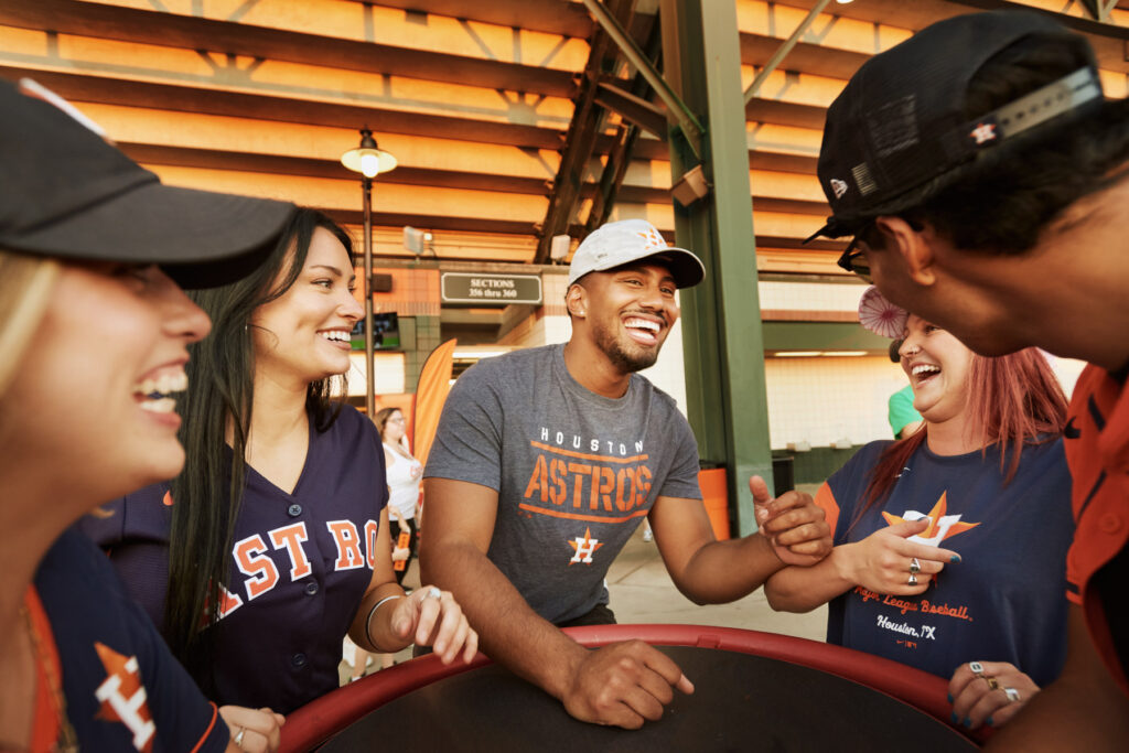 A group of five people, wearing Houston Astros shirts and caps, smile and laugh together inside a stadium. They appear to be enjoying themselves before or during a baseball game.