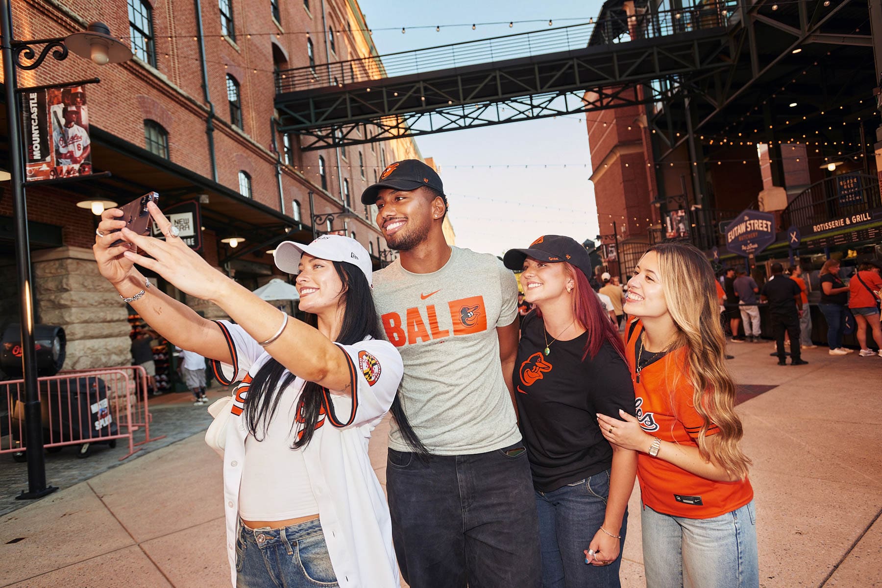 Four young adults wearing Baltimore Orioles gear smile and pose for a selfie outside a baseball stadium, with brick buildings, string lights, and other fans in the background.