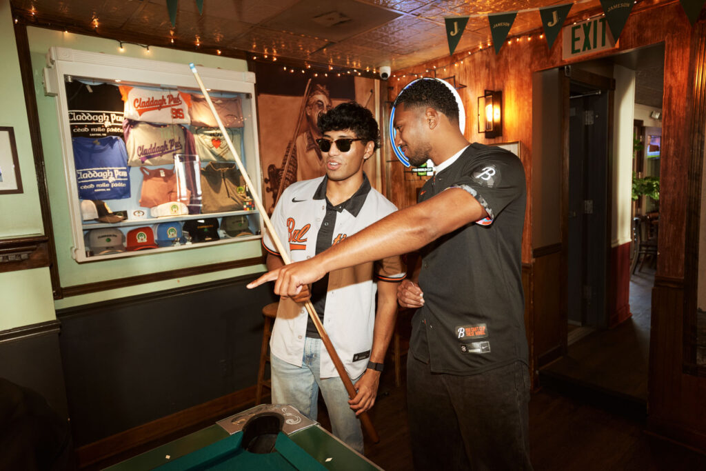 Two young men stand by a pool table in a bar. One wears sunglasses and holds a cue stick, while the other points, possibly giving advice. The bar has dim lighting, decorations, and wall art in the background.