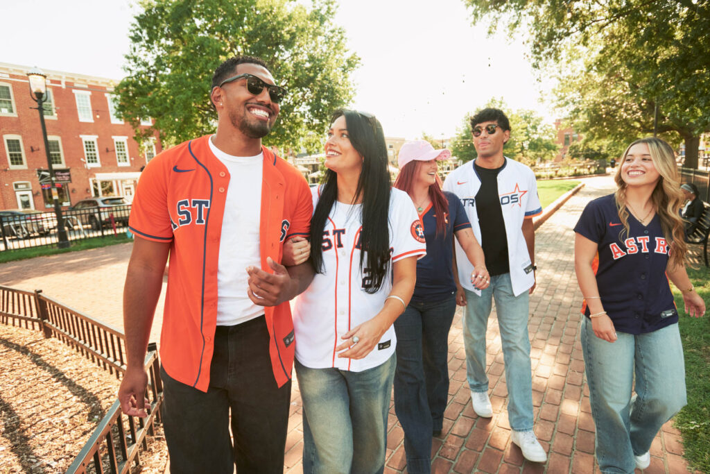 Five young adults walk together in a park, smiling and wearing various Houston Astros baseball jerseys and casual clothes, with trees and historic brick buildings in the background.
