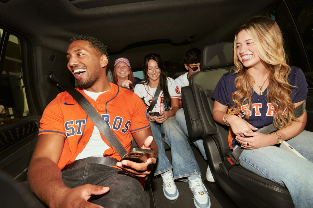 Five young adults in Astros jerseys sit in a car, laughing and smiling. Two are in the front row, three in the back; some are looking at phones. The atmosphere is cheerful and friendly.