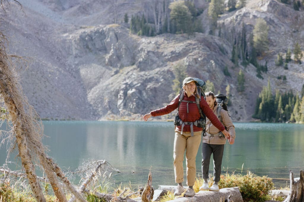 Two people with backpacks balance on rocks by a lake in a mountainous forest area. One person walks ahead with arms outstretched, while the other follows closely behind. Pines and rocky hills are in the background.