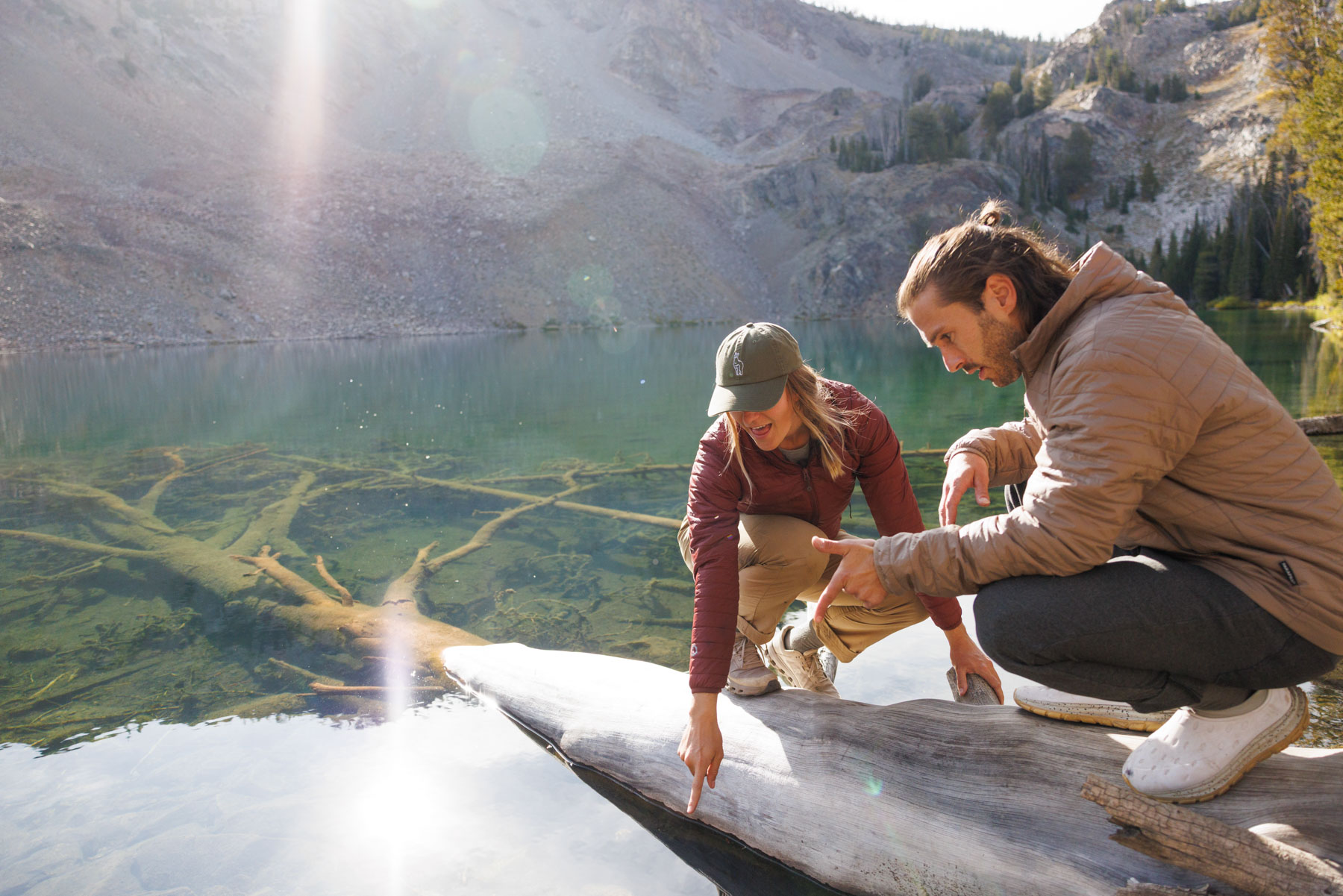 Two people crouch on a large log by a clear mountain lake, pointing and looking at something in the water. Trees and rocky hills are reflected in the calm lake behind them.