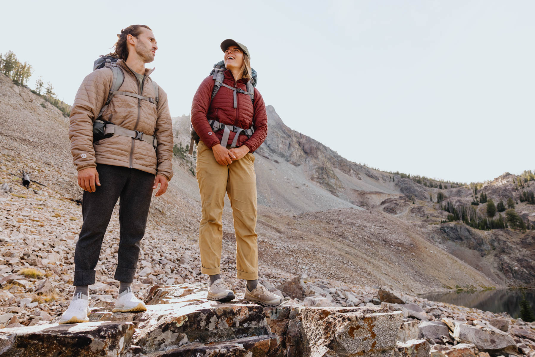 Two people wearing outdoor jackets and backpacks stand on rocky terrain, looking at each other and smiling, with a mountainous landscape and sparse trees in the background under a clear sky.