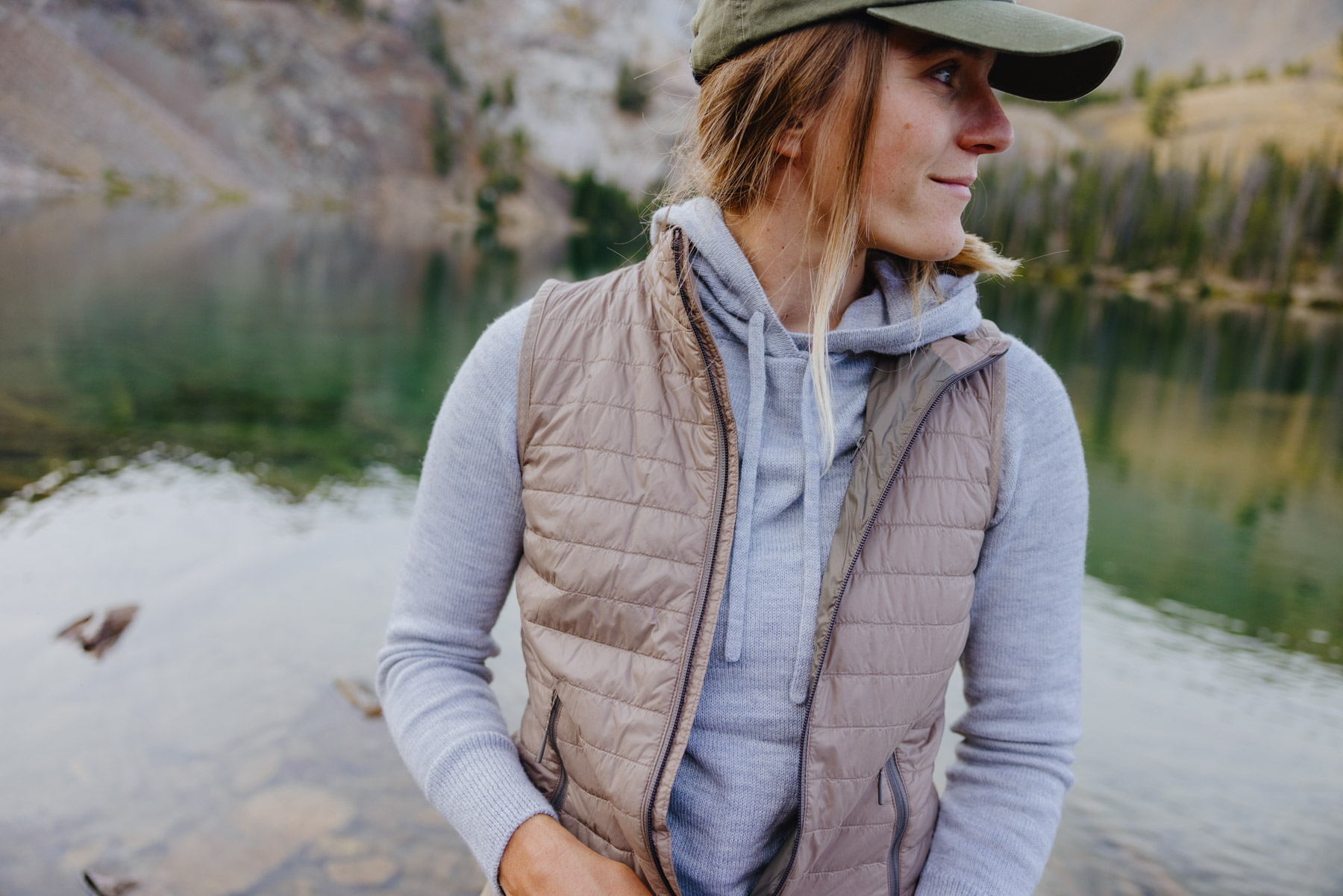 A person wearing a gray hoodie, beige quilted vest, and green cap stands by a clear mountain lake, looking to the side. Pine trees and rocky slopes are visible in the background.