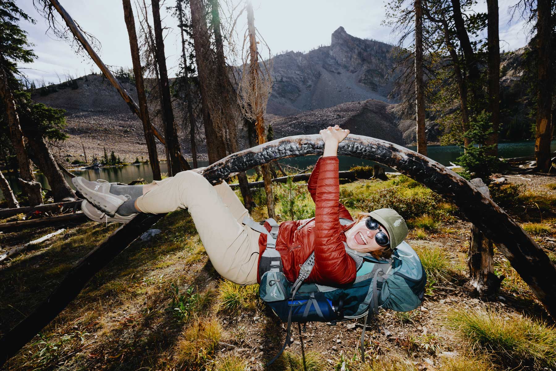 A person in outdoor gear and sunglasses hangs playfully from a curved tree branch, smiling, with a large backpack on the ground and mountains, trees, and a lake in the background.