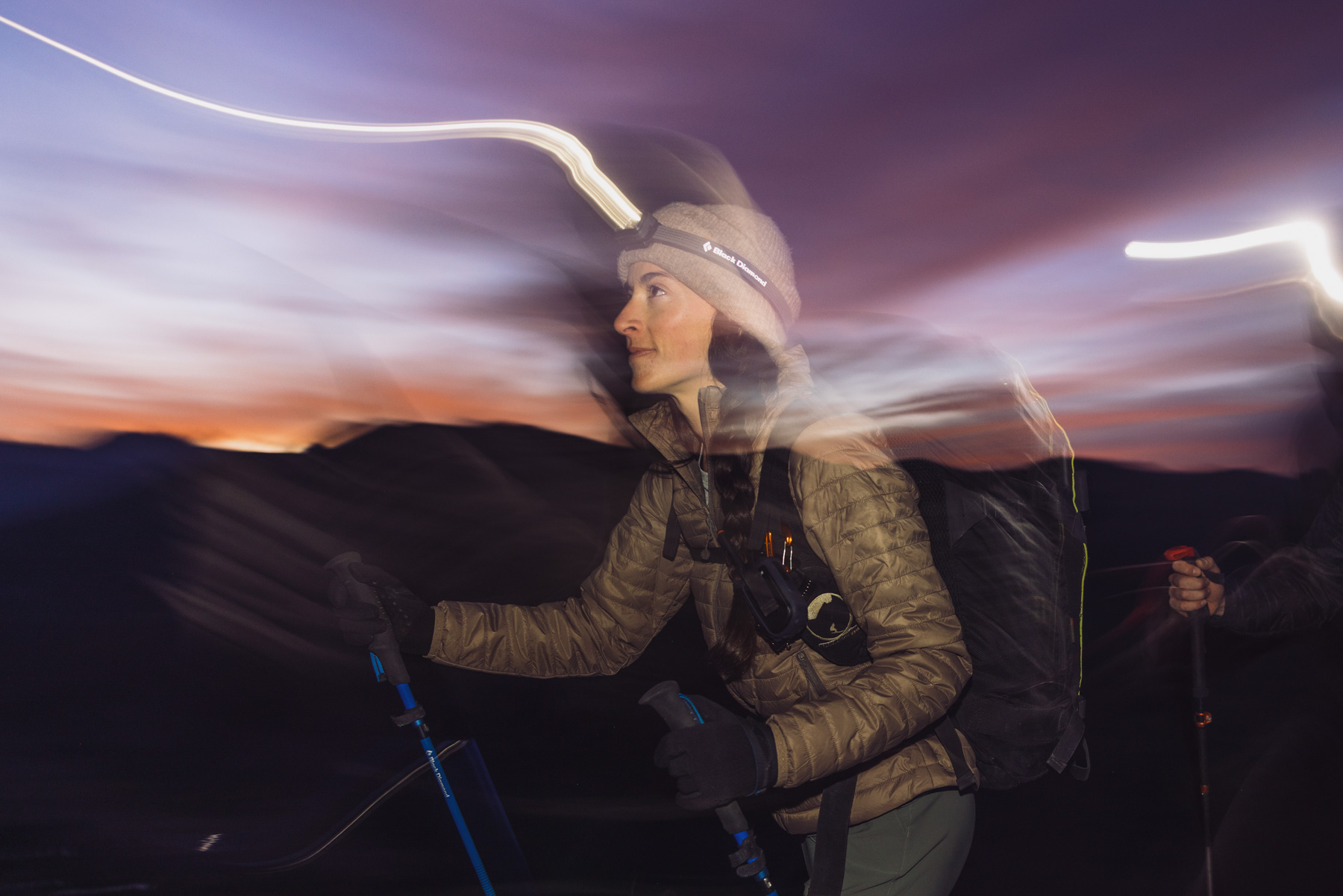 A woman hiking at dusk with trekking poles and a backpack. Light trails swirl around her, suggesting movement, as the sky glows with deep purple and orange tones in the background.