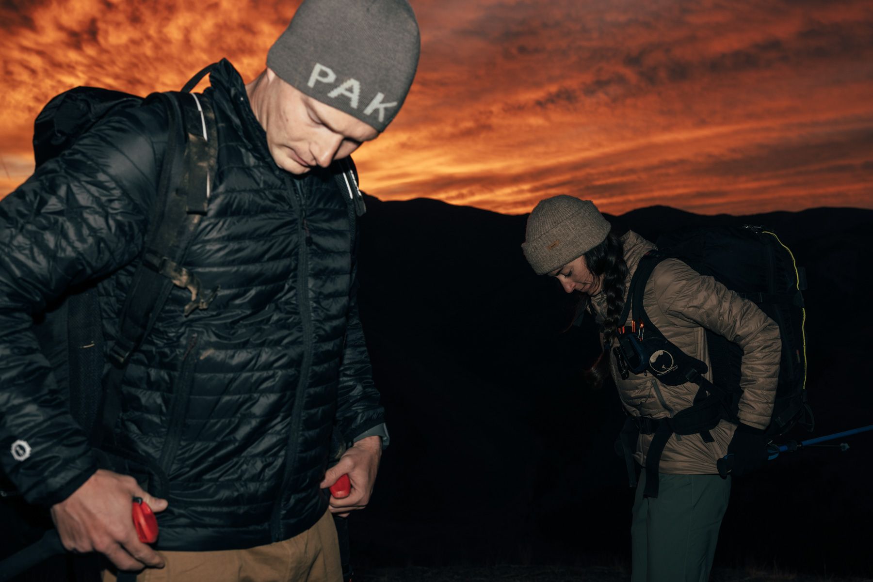 Two hikers wearing beanies and backpacks adjust their gear at dusk, with an orange and red sunset sky and dark mountains in the background.