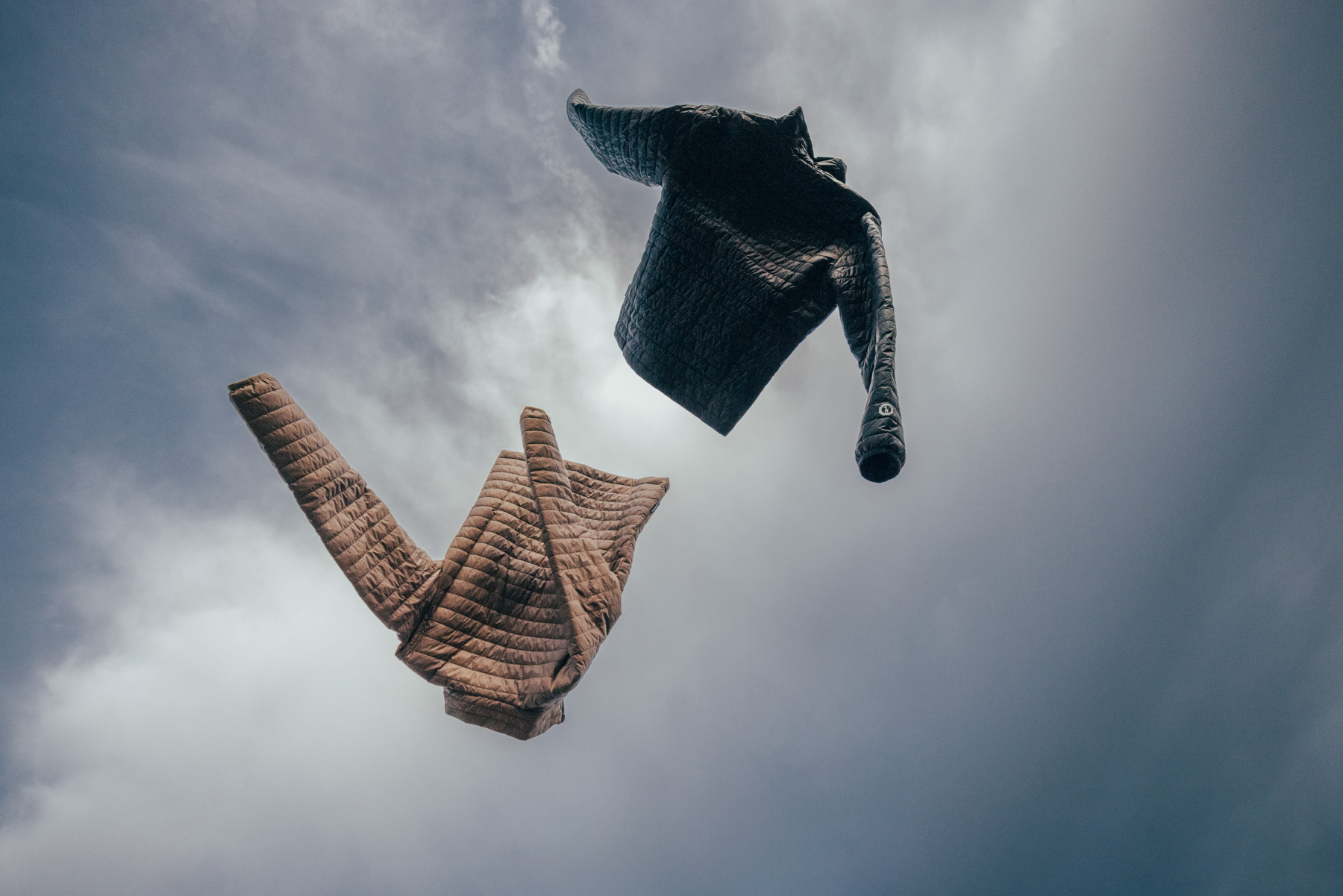 Two padded jackets, one brown and one black, appear to be floating or flying against a cloudy, overcast sky. The image is taken from a low angle, creating a surreal and dynamic scene.