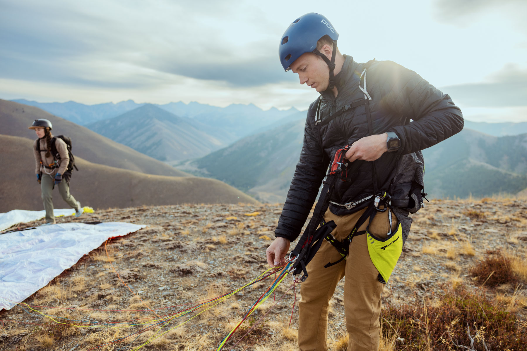 A person wearing a helmet and harness prepares colorful paragliding lines on a mountain slope, with another person and a paraglider in the background, surrounded by distant mountains under a cloudy sky.