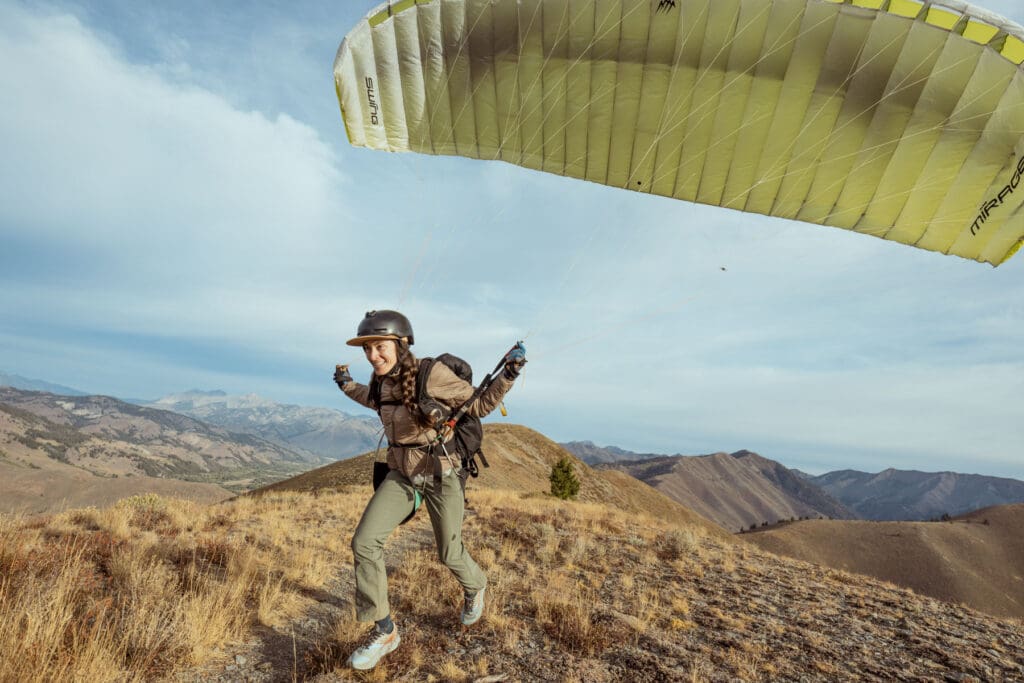 A person wearing a helmet and harness runs on a grassy hilltop, preparing to launch a yellow paraglider, with mountains and a cloudy sky in the background.