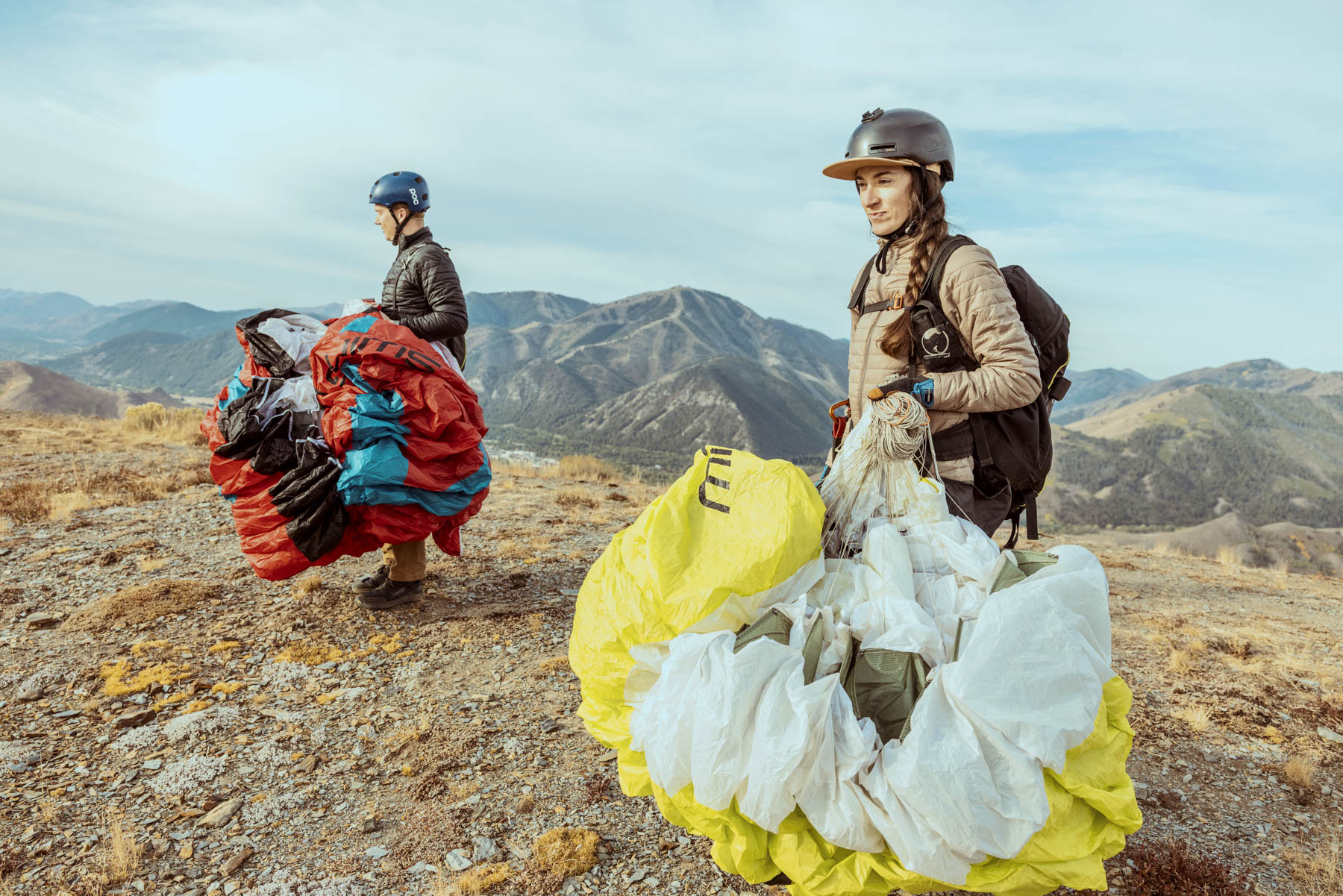 Two people wearing helmets and outdoor gear stand on a rocky mountain slope, each holding a packed parachute with colorful fabric, with mountains and a cloudy sky in the background.