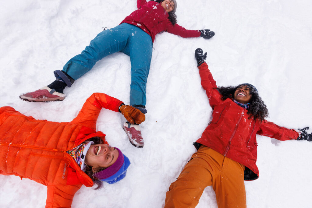 Three people in colorful winter clothes lie on their backs in the snow, smiling and laughing with arms and legs outstretched, enjoying a fun moment outdoors together.
