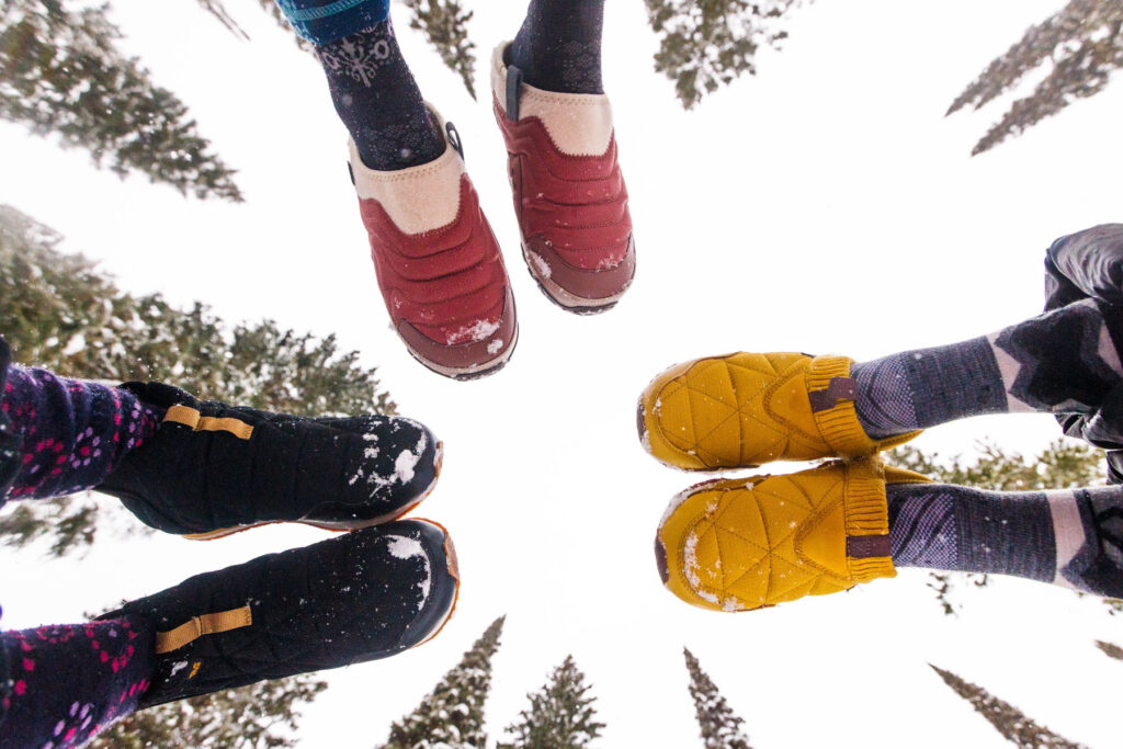 Four people wearing colorful winter boots and socks stand in a circle on snowy ground, with tall pine trees and a bright sky in the background, viewed from below looking up.