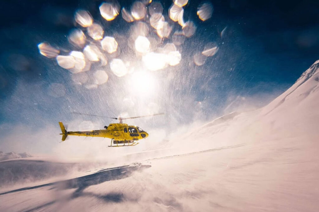A yellow helicopter flies low over a snowy mountain slope under a bright sun, with snow swirling in the air and lens flares visible in the sky.