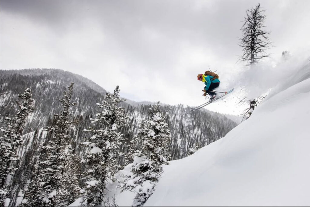 A skier in bright gear leaps off a snowy slope, surrounded by snow-covered pine trees and mountains under a cloudy sky.