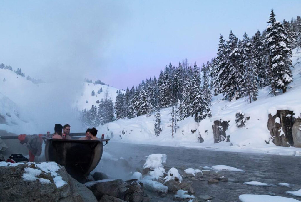 Three people relax in a steaming hot tub beside a snowy river, surrounded by snow-covered trees and mountains under a pale sky.