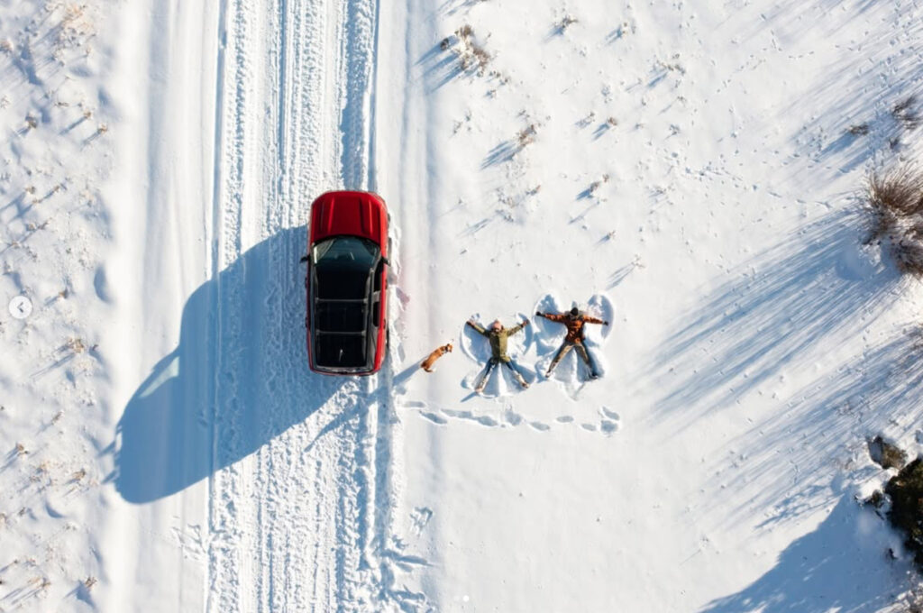 A red SUV is parked on a snowy road. Two people are lying in the snow making snow angels, and a dog stands nearby. The scene is viewed from above.