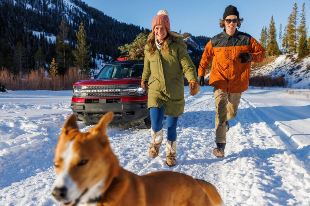 A woman and man in winter clothes run joyfully on a snowy road with a dog in the foreground. A red SUV with a Christmas tree on top is parked behind them. Snowy mountains and pine trees surround the scene.