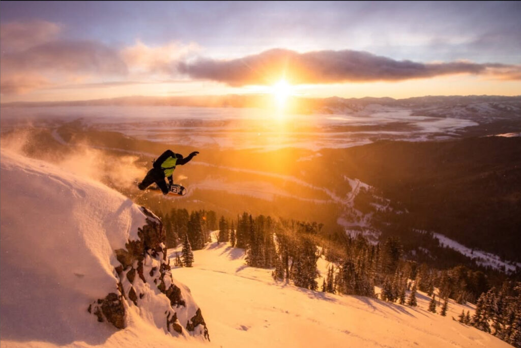 A snowboarder in mid-air leaps off a snowy mountain cliff at sunset, with golden sunlight illuminating the snow and a vast landscape of trees and mountains in the background.
