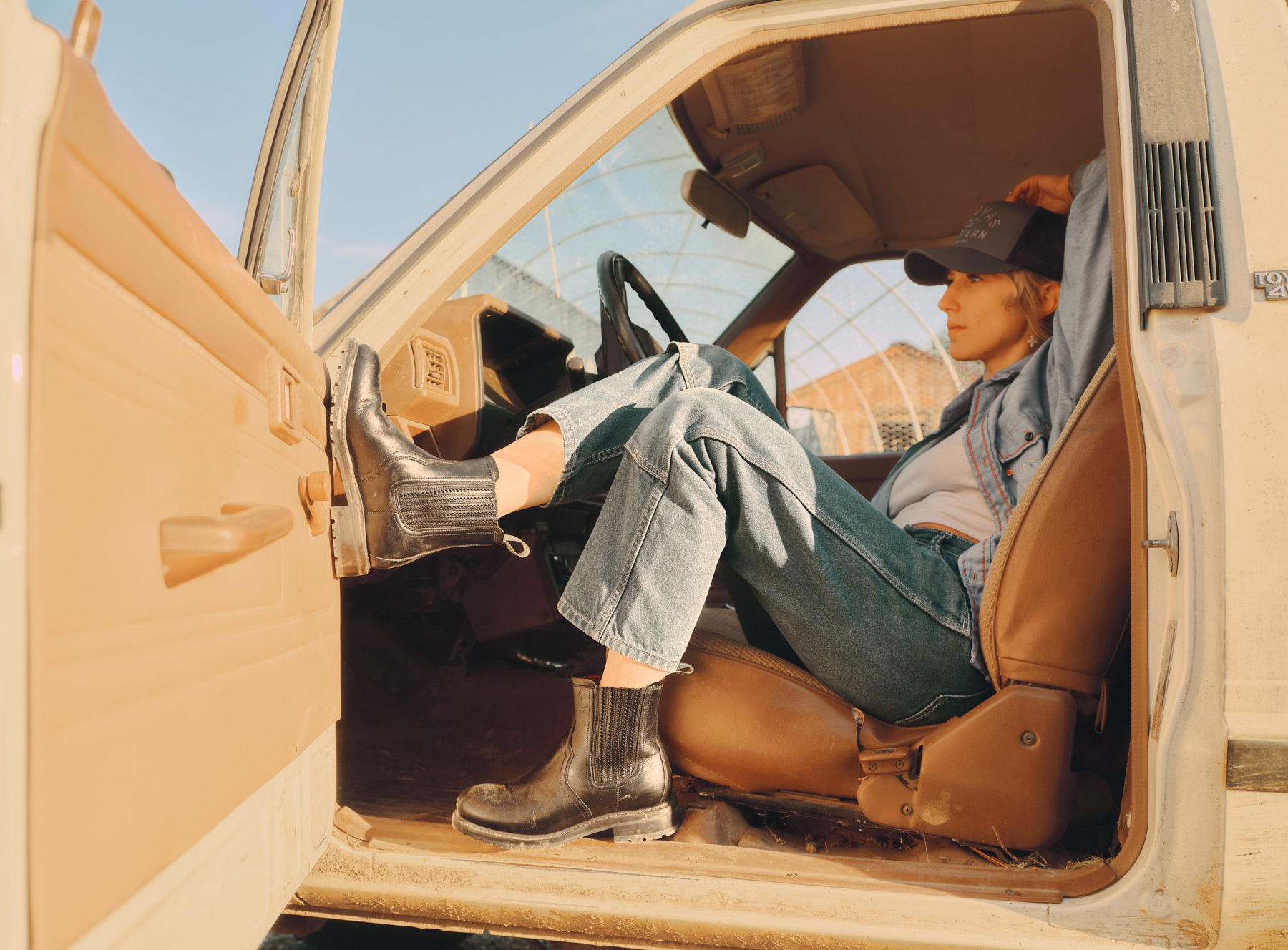 A person wearing jeans, a denim shirt, and black boots relaxes in the drivers seat of an old beige truck with the door open, one foot on the dashboard, looking out into the distance on a sunny day.