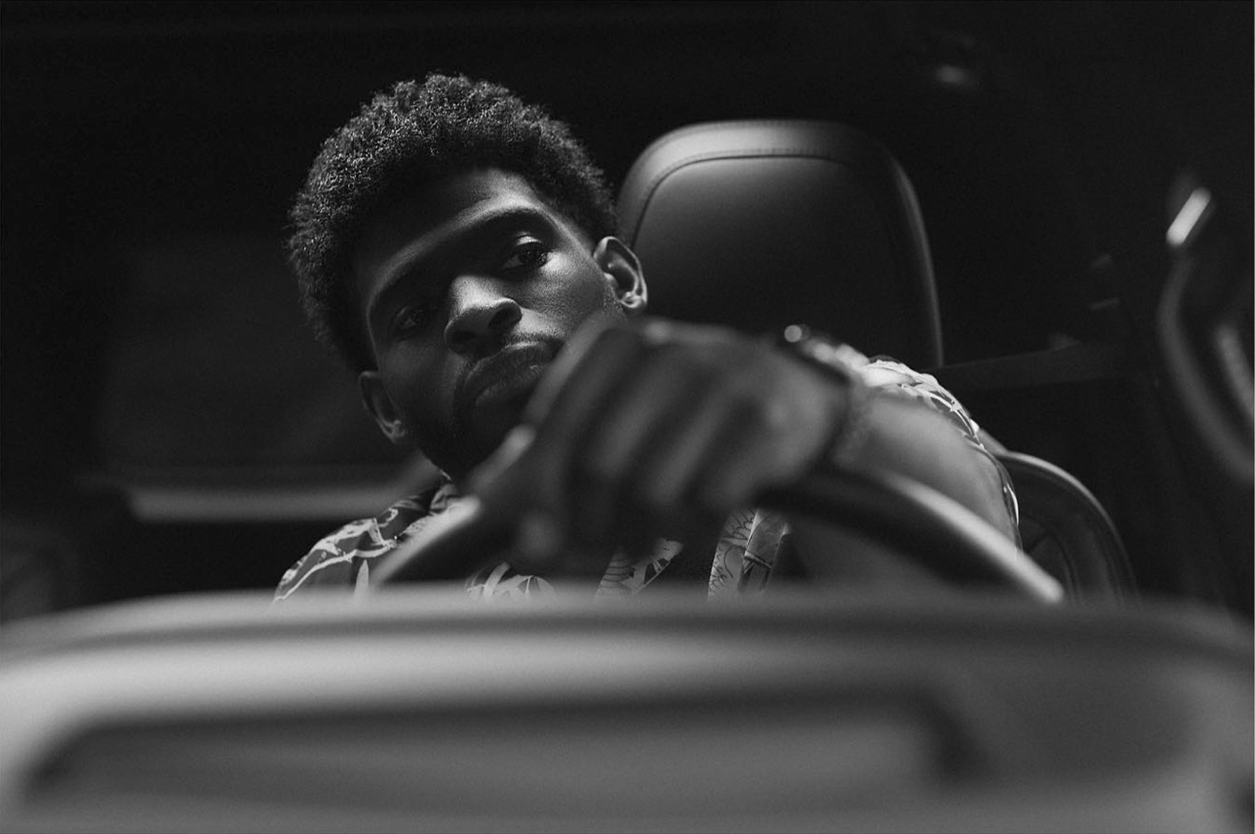 Black and white photo of a man sitting in the drivers seat of a car, viewed from the dashboard. He has one hand on the steering wheel and looks ahead with a serious expression.