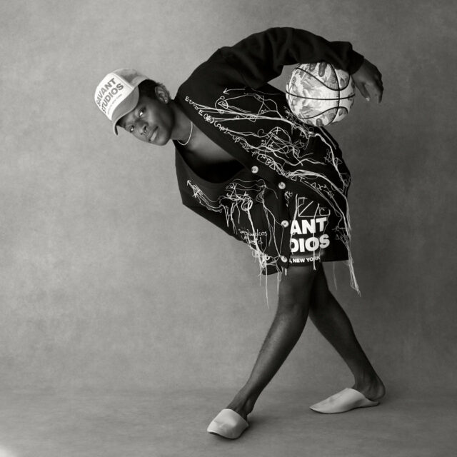 A person in a cap, oversized jacket, and shorts poses with a basketball balanced on their back. The outfit features abstract line art and text. They stand barefoot against a plain, textured background in a dynamic, fashion-forward stance.