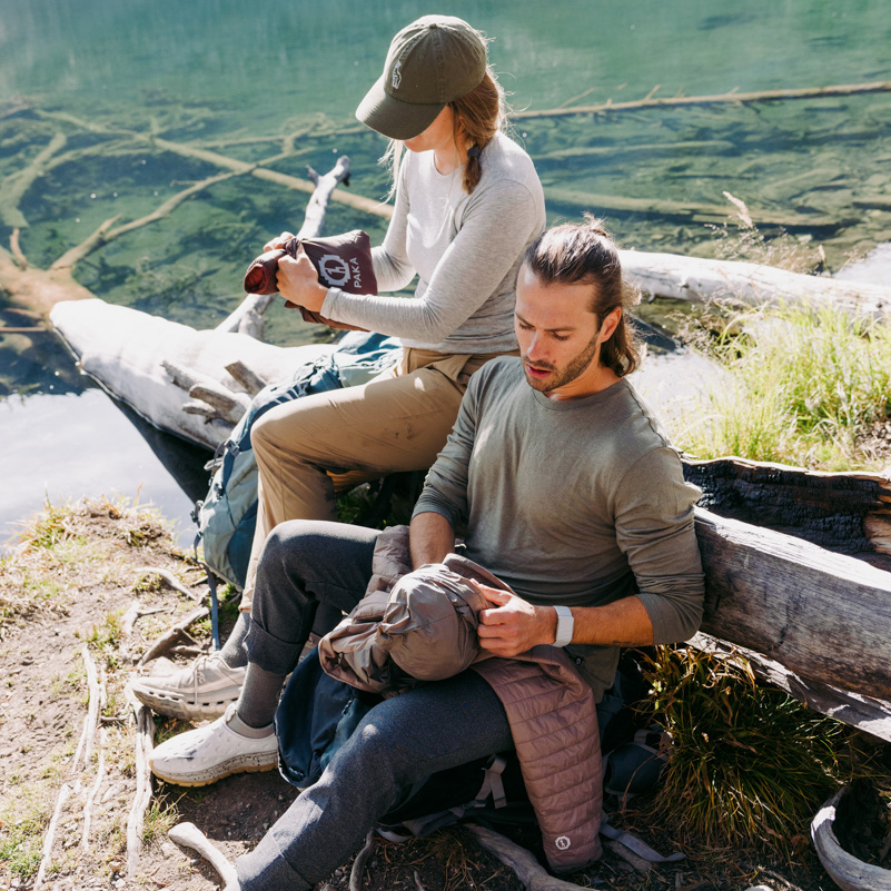 Two people wearing casual outdoor clothes sit on a log by a lakeshore, unrolling jackets or clothing from their bags. The water is calm, and fallen logs are visible in the lake behind them.