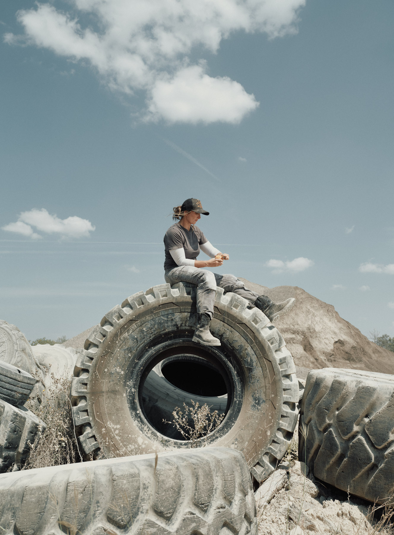 A person wearing a cap and work clothes sits on top of a large, weathered tire in an outdoor, industrial area with piles of dirt and more tires under a cloudy blue sky.