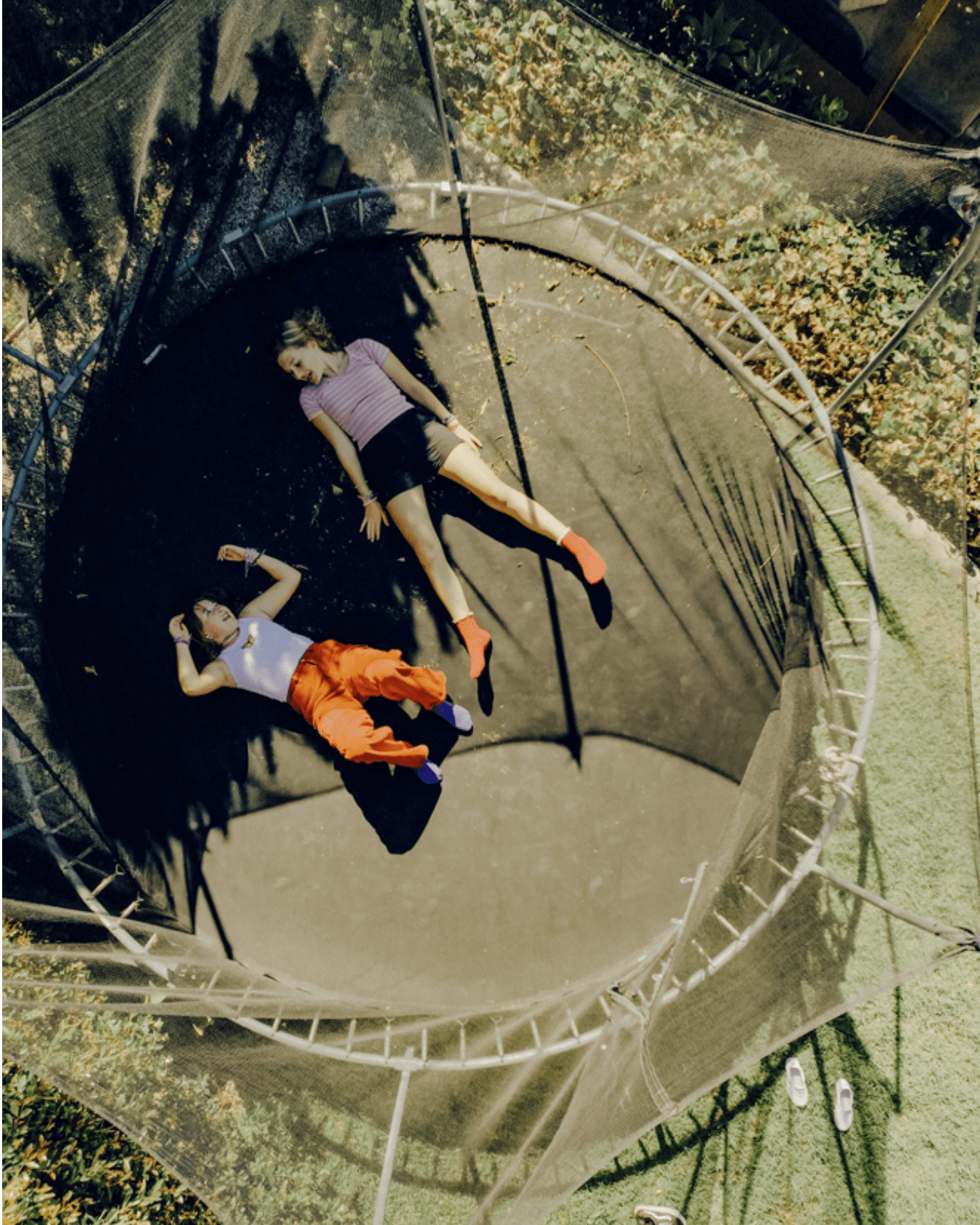 Two people lie on their backs on a trampoline outdoors, relaxing. One holds a phone, both wear socks with their shoes left nearby on the grass. The trampoline is surrounded by a safety net and dappled sunlight.
