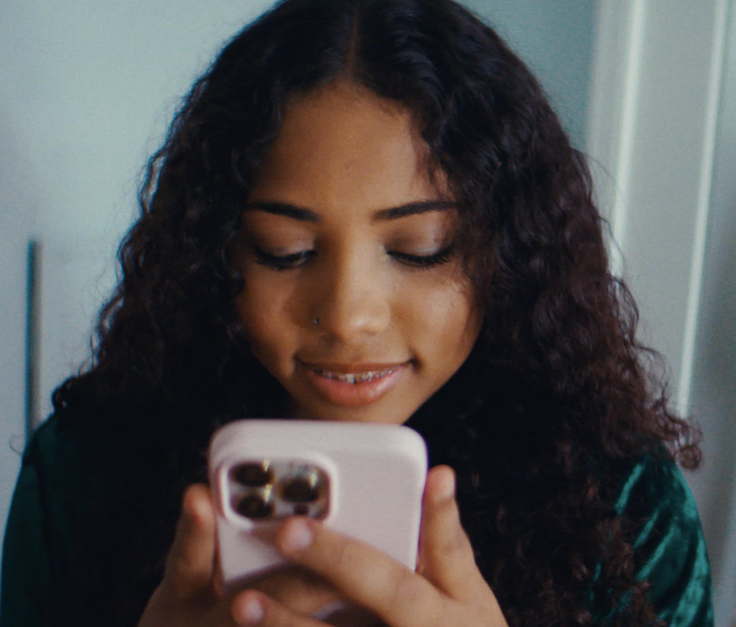 A person with curly hair smiles while looking down at a smartphone with a pink case in their hands.