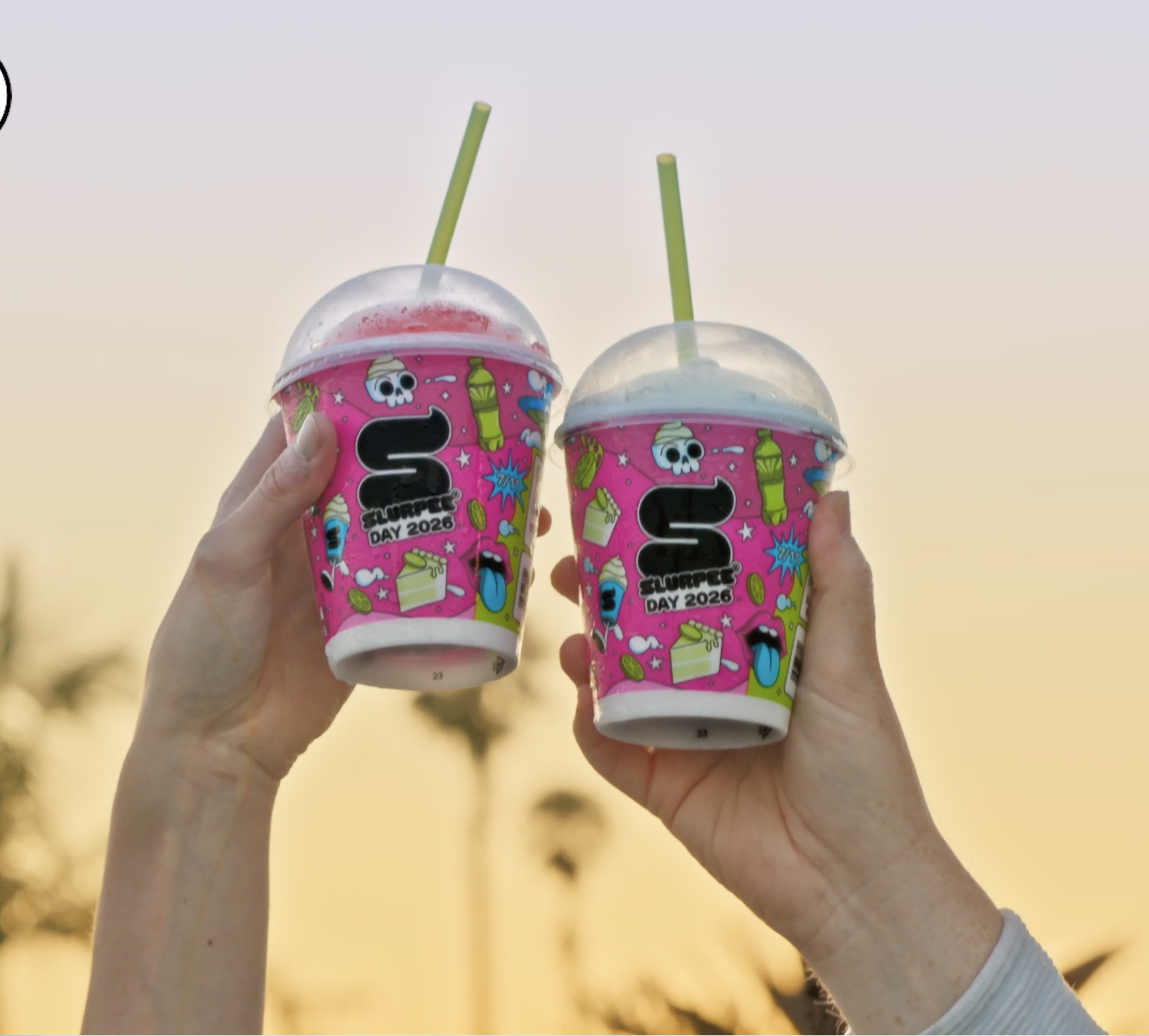 Two hands hold up pink Slurpee cups with green straws and colorful designs. The cups each feature Slurpee Day 2023. Palm trees are blurred in the sunset background.