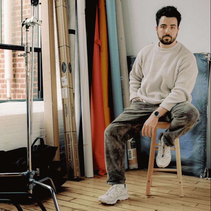 A man in a beige sweater and grey pants sits on a wooden stool in a studio with photography equipment and rolls of colored paper in the background.
